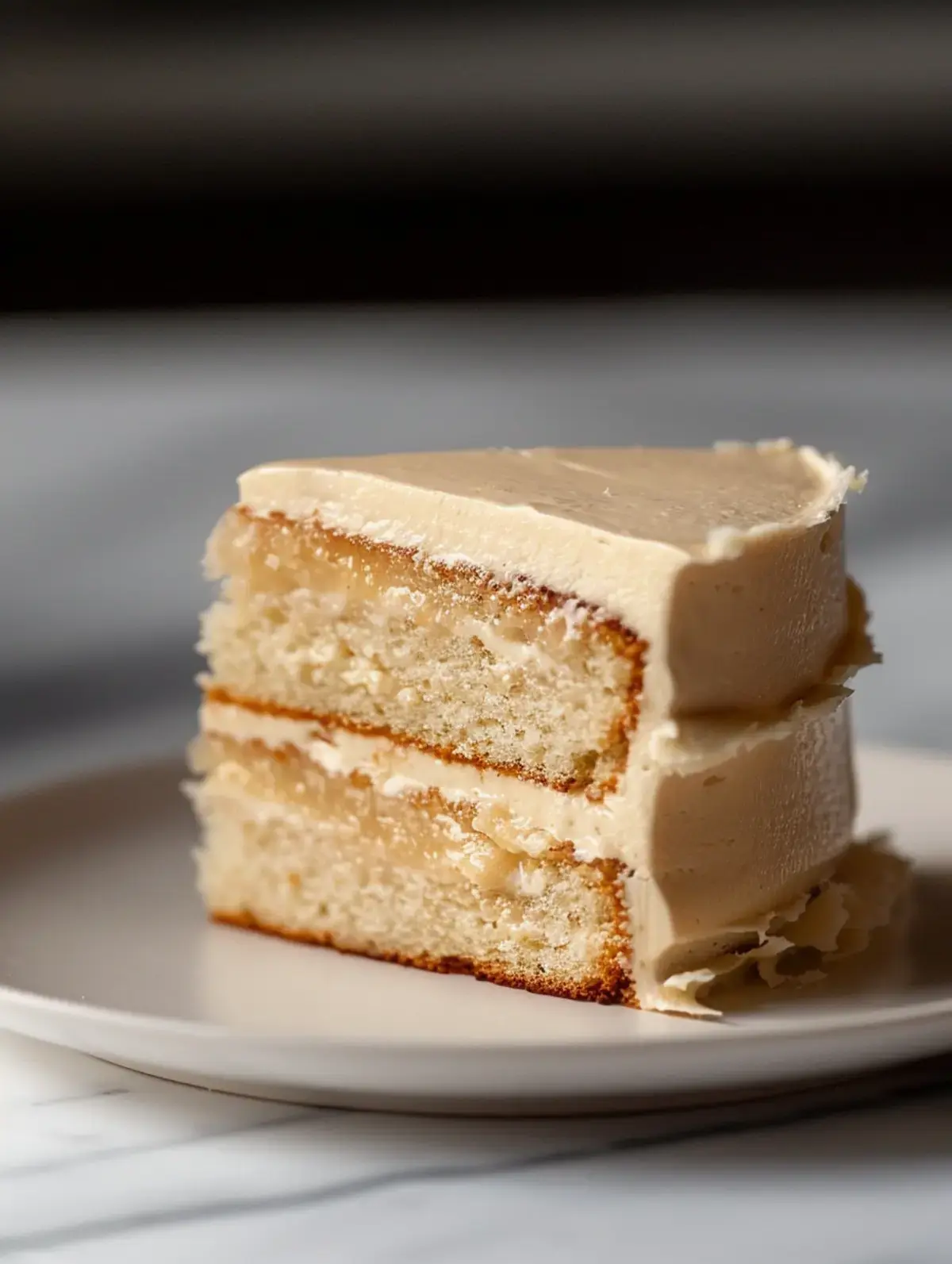 Slice of white chocolate layer cake on a ceramic plate, showing two moist sponge layers with a thick filling and soft frosting exterior.