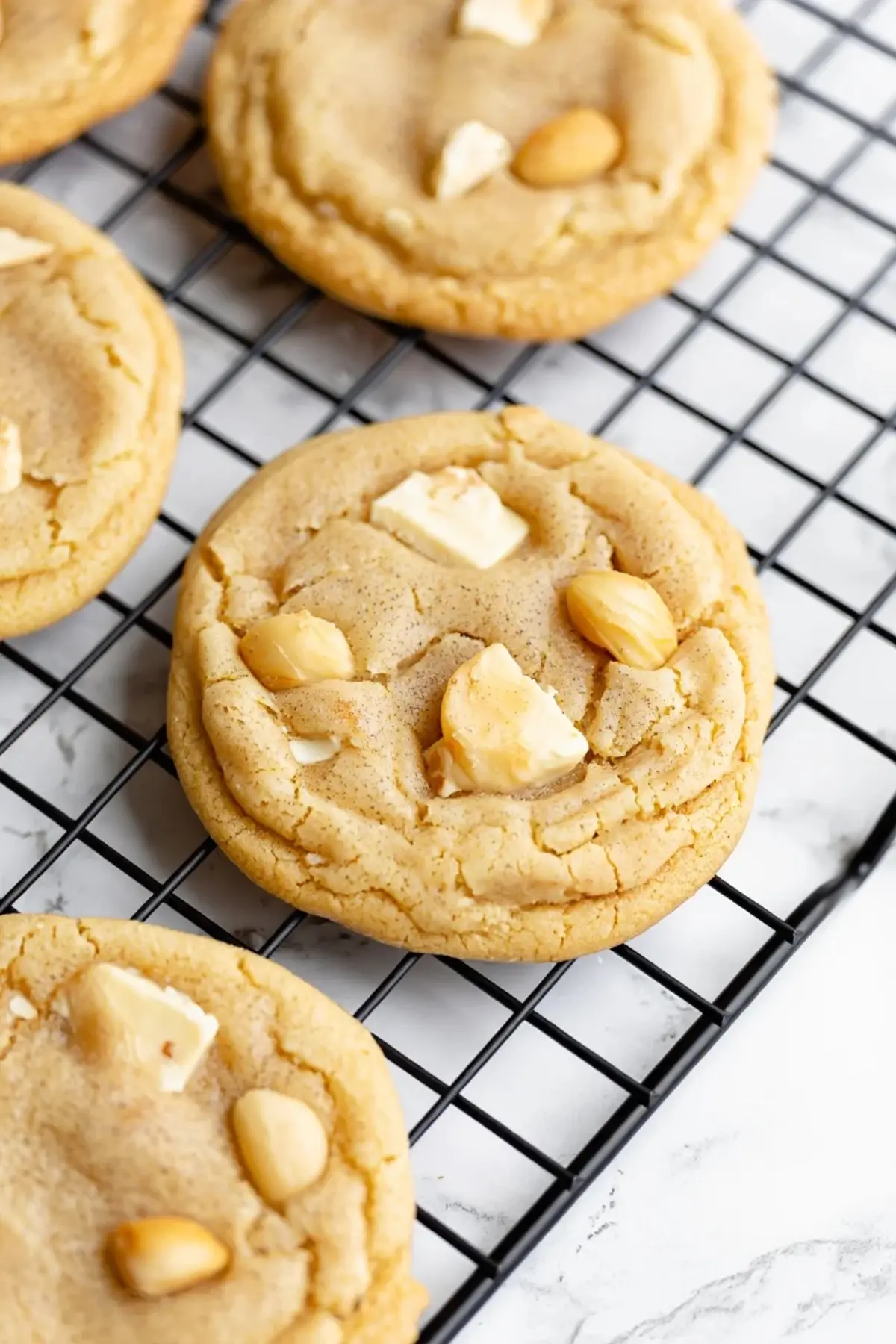 Close-up of golden brown white chocolate macadamia nut cookies on a black cooling rack, with visible chunks of chocolate and nuts on a marble background.