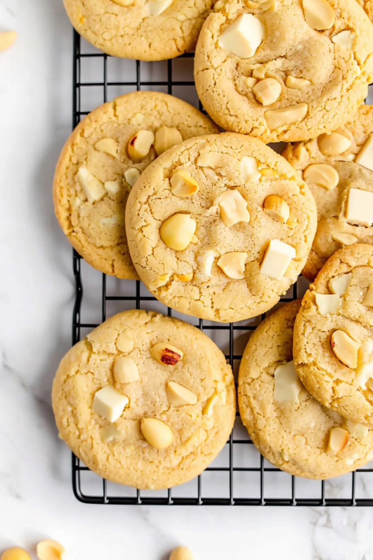 Cooling rack filled with golden white chocolate macadamia nut cookies topped with chunks of chocolate and whole nuts on a marble surface.