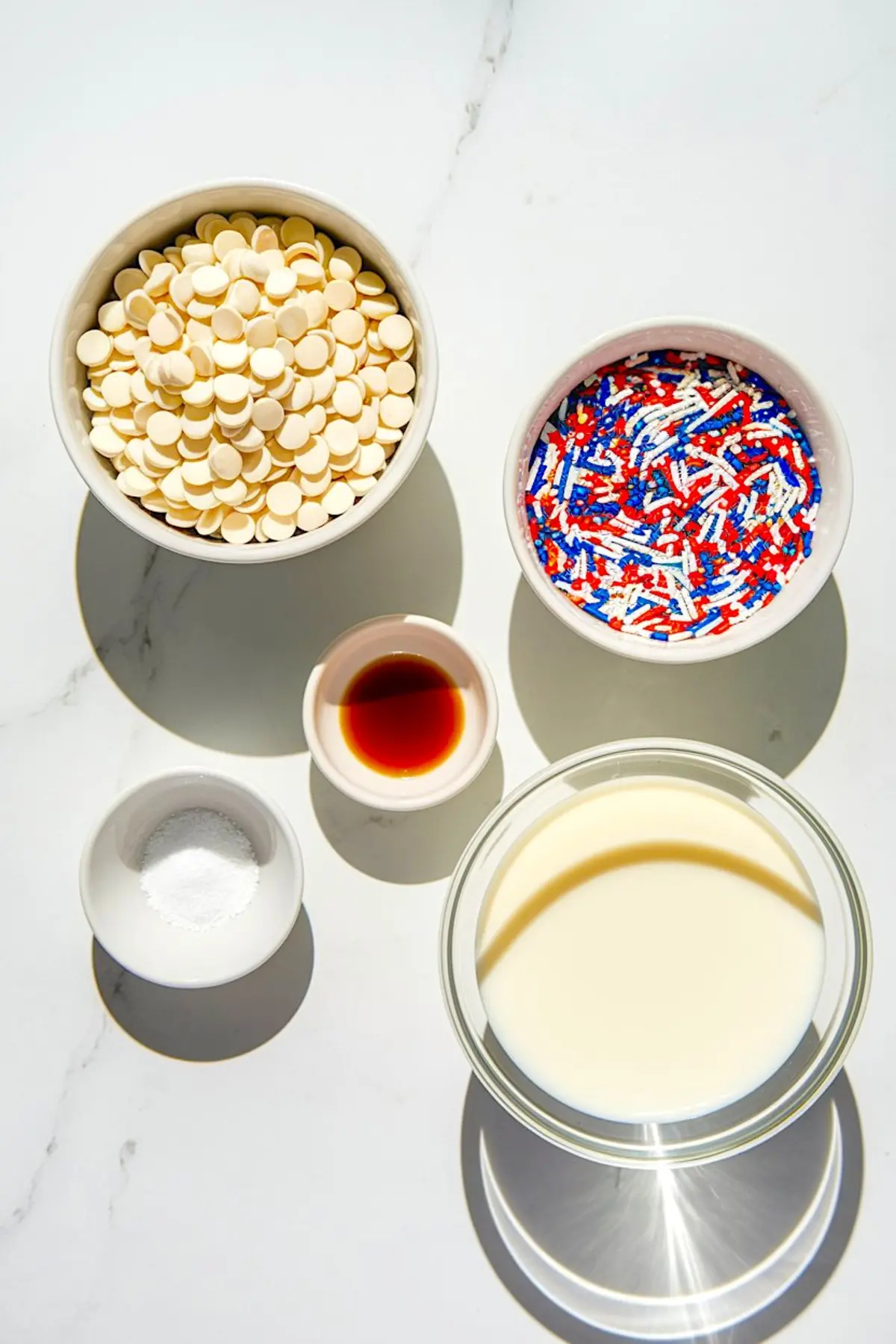 Flat lay of fudge ingredients on a white surface, including bowls of white chocolate chips, red-white-blue sprinkles, vanilla extract, sugar, and condensed milk, arranged for easy no-bake recipe prep.