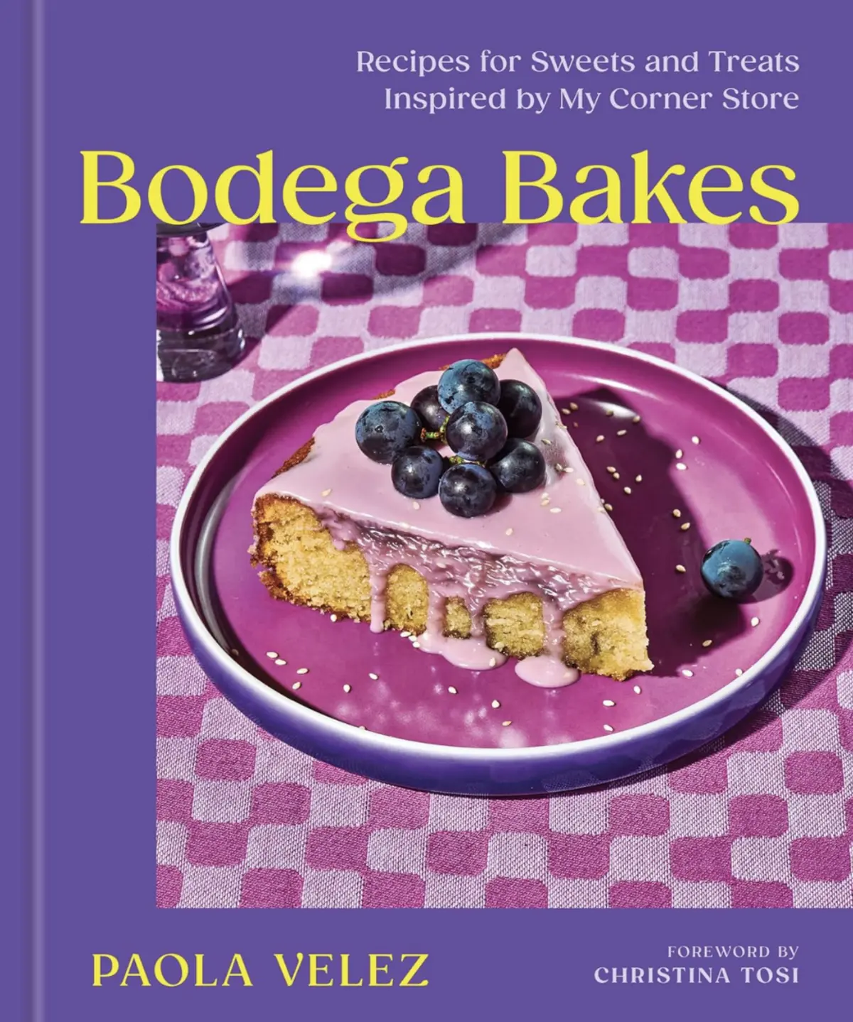 “Bodega Bakes” by Paola Velez features a vibrant purple-themed cover with a slice of yellow cake topped with pink icing, fresh blueberries, and sesame seeds, served on a magenta plate over a checkered cloth.
