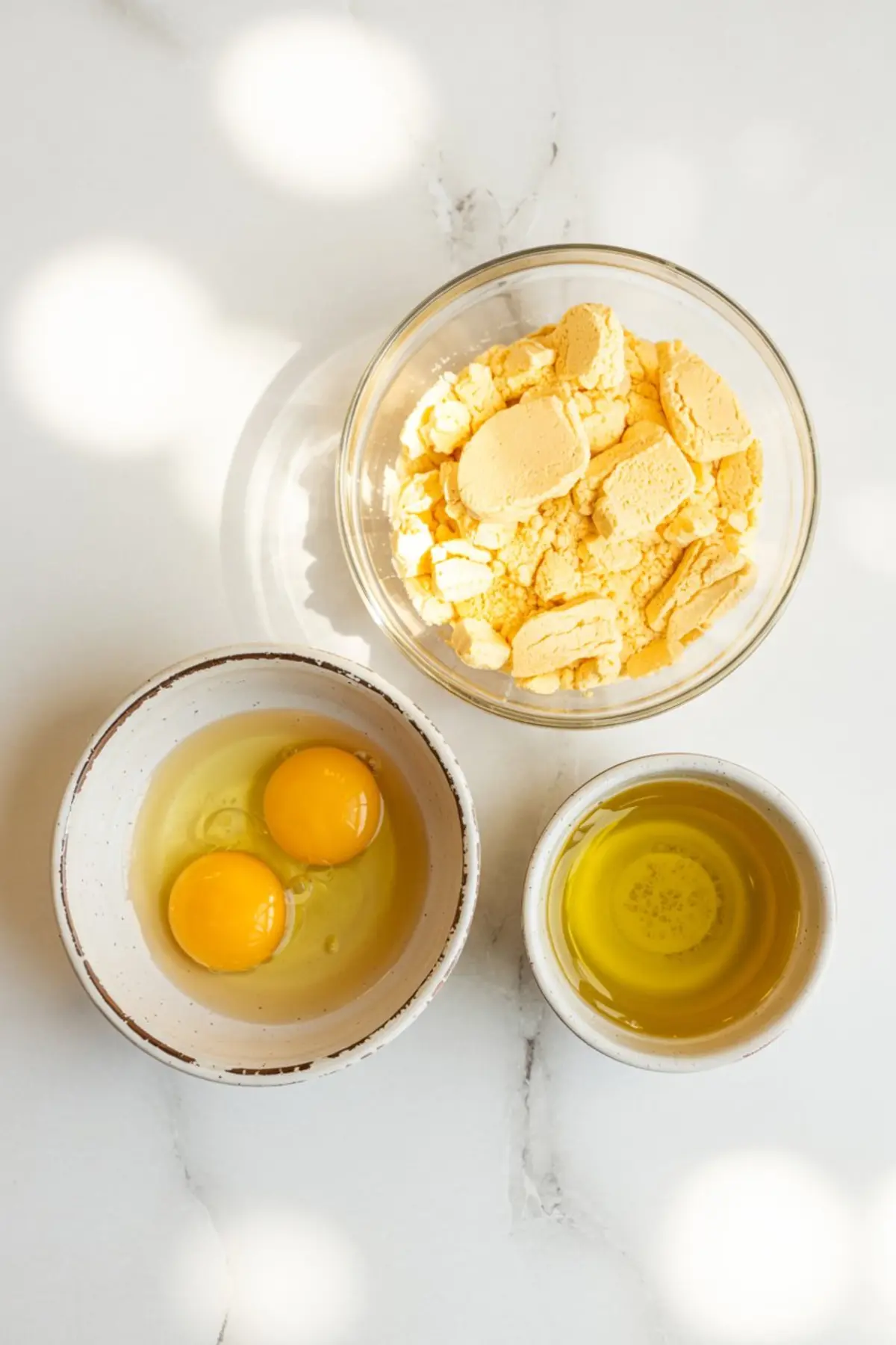 Flat lay of three bowls showing separated ingredients for cake mix cookies: yellow cake mix, raw eggs, and vegetable oil, essential for simple cake box cookies.