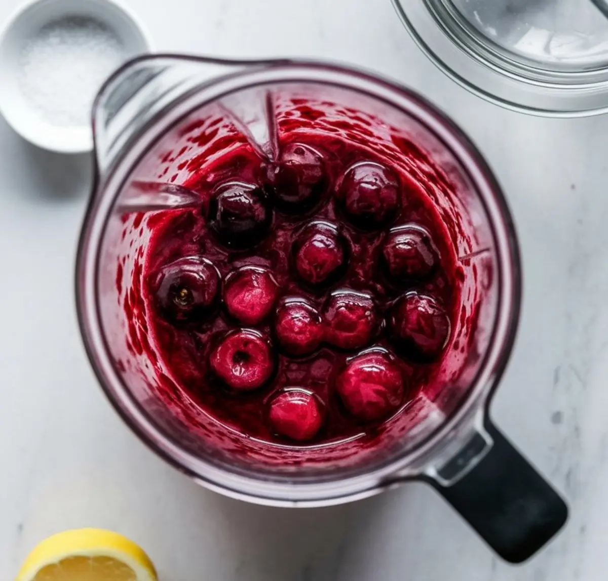 Top view of a blender filled with partially blended fresh cherries in a thick red purée, surrounded by ingredients for making cherry sorbet.