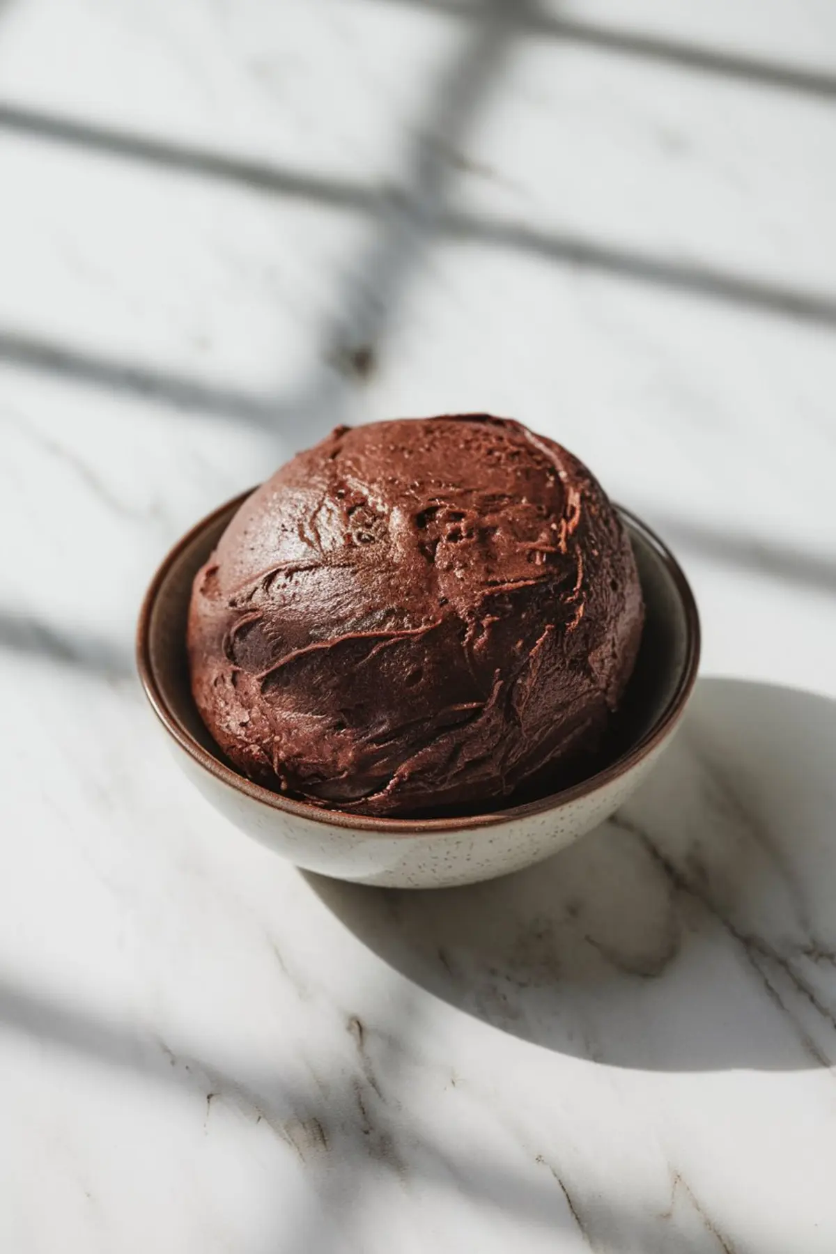 Close-up of a bowl of chocolate cookie dough with a smooth texture and rich brown color, sitting on a marble countertop with natural lighting.