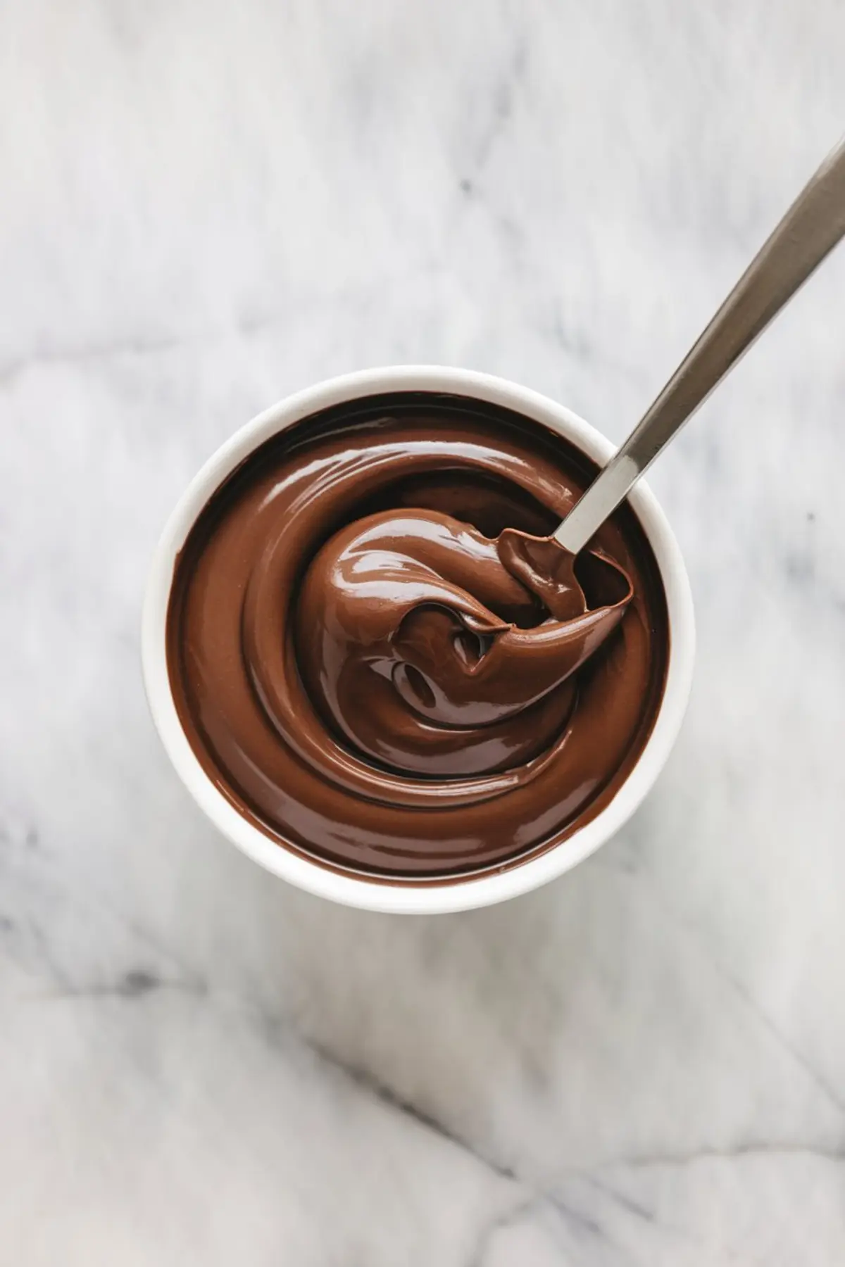 Top view of a small white bowl filled with glossy chocolate ganache, swirled smoothly with a spoon resting inside on a light marble surface.