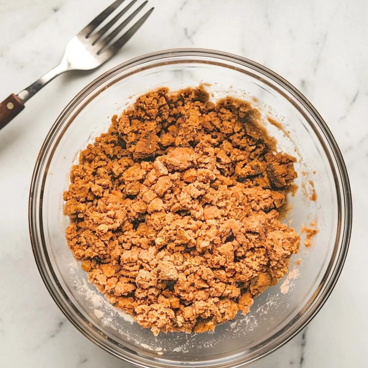 Glass bowl of cinnamon streusel crumble topping with a coarse texture, sitting on a marble surface next to a metal fork, perfect for coffee cake or crumb bars.
