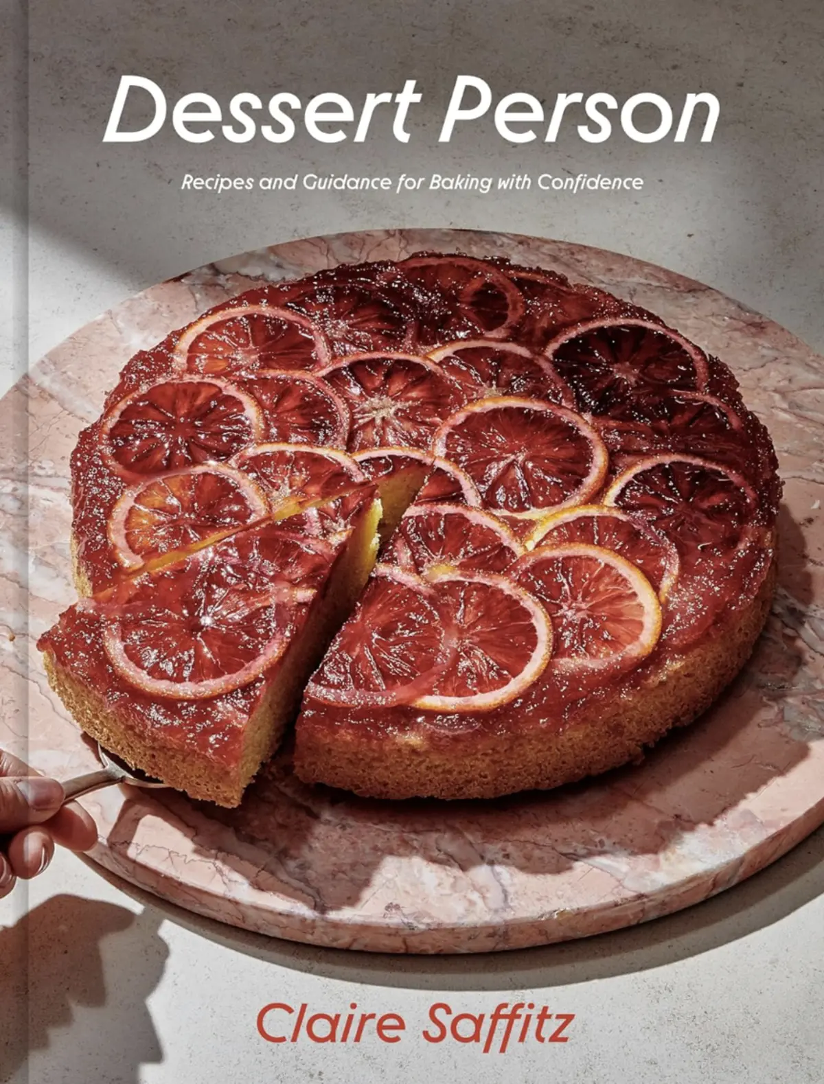 Cookbook cover for “Dessert Person” by Claire Saffitz showing a blood orange upside-down cake sliced on a marble platter, with caramelized citrus topping glistening under natural light.