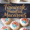 Macarons with firework-style painted tops in bold red and blue strokes, filled with vanilla buttercream and styled on a marble background for a patriotic or celebratory baking theme.