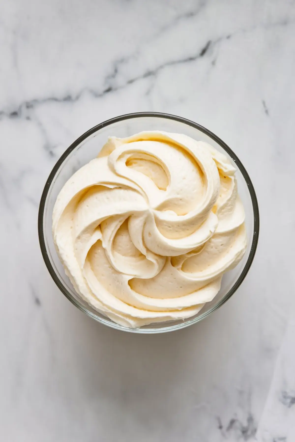 Glass bowl filled with smooth vanilla buttercream swirled into soft peaks, displayed on a marble surface for cake or macaron filling inspiration.
