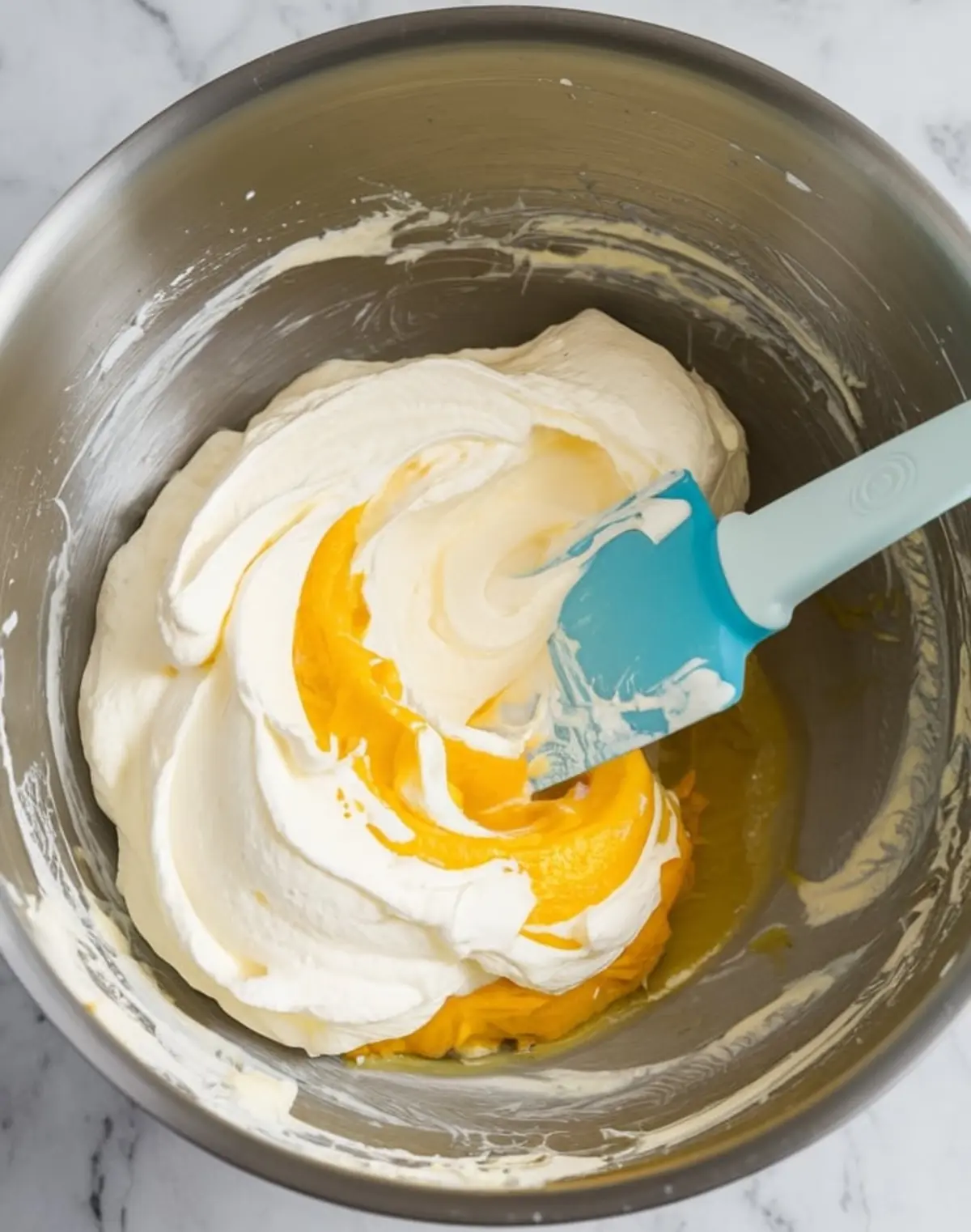 Close-up of whipped cream and lemon curd swirled together in a stainless steel mixing bowl with a spatula, mid-step in preparing frozen lemon soufflé batter.
