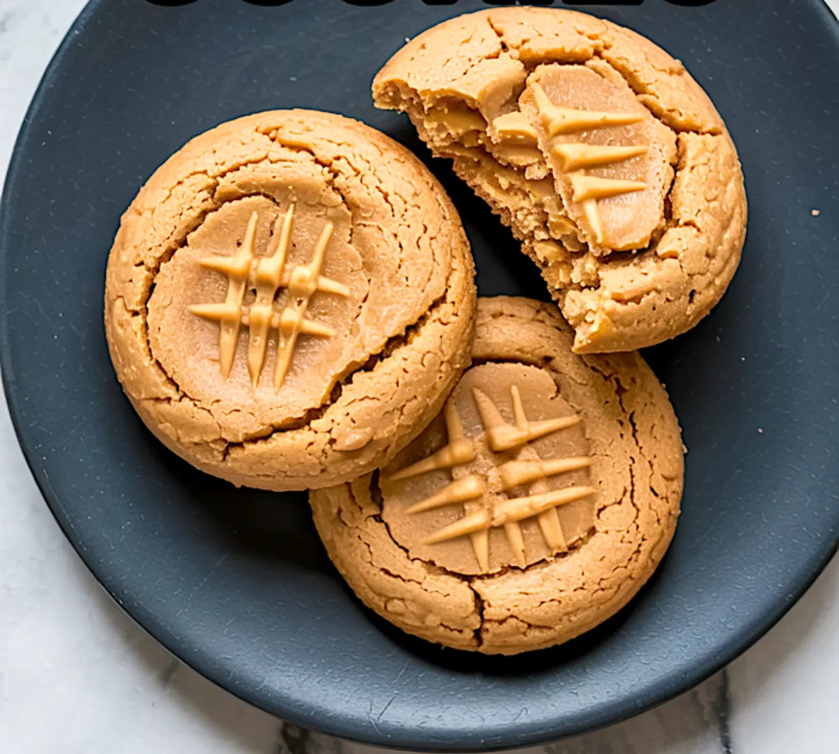 Close-up of three keto peanut butter cookies on a dark plate, with one cookie broken in half to show the soft interior and crosshatch design on top.