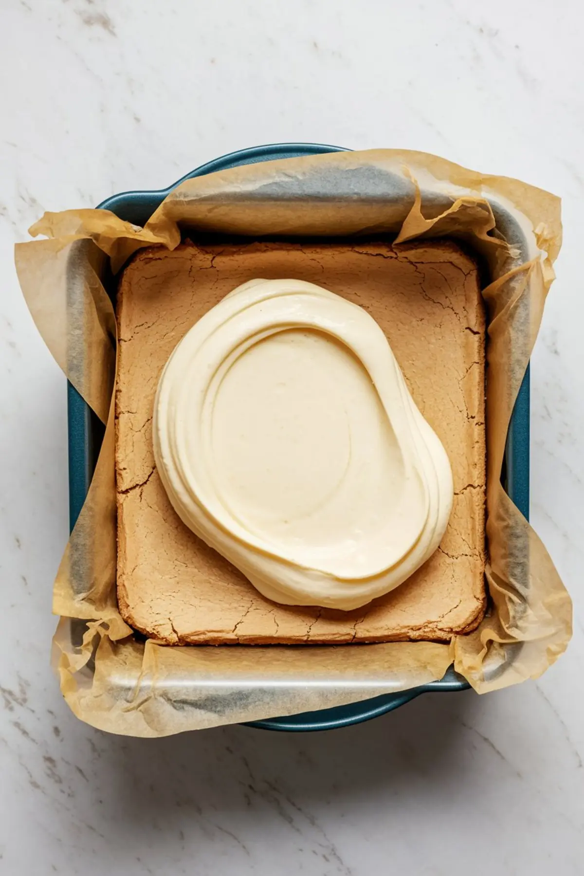 Overhead image of a parchment-lined square baking pan with golden brown cookie crust topped with a thick swirl of creamy white cheesecake filling ready for layering.