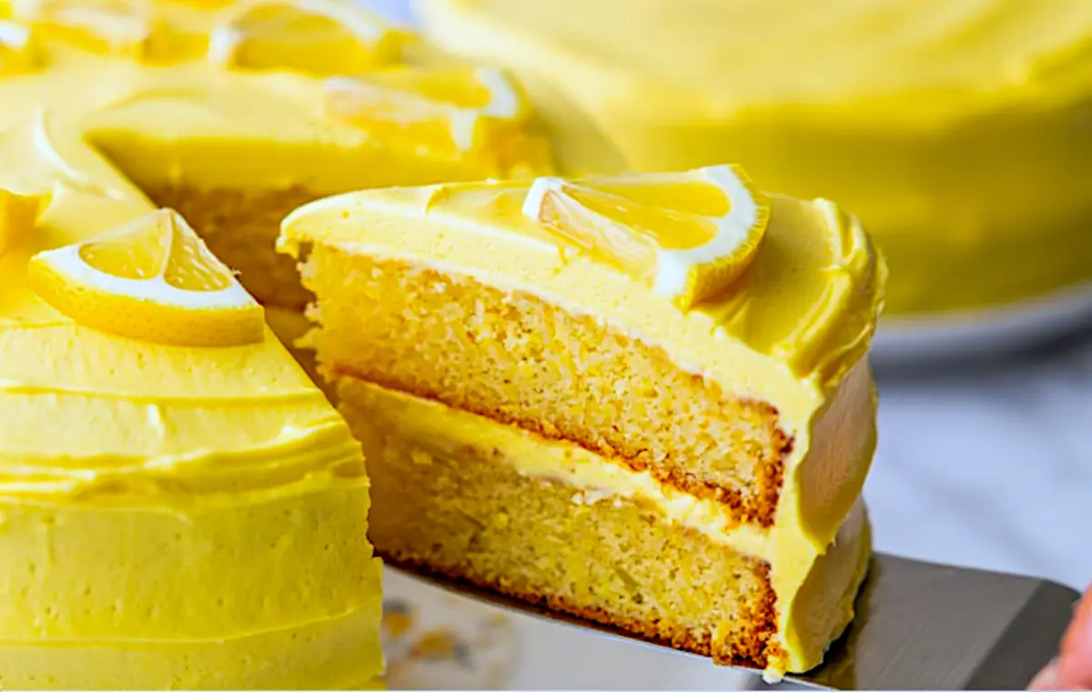 Close-up of a lemon cake slice being served, showing moist layers and thick yellow frosting topped with a lemon wedge.