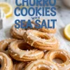 Ring-shaped lemon churro cookies topped with vanilla glaze and sea salt flakes, arranged on parchment paper with lemon slices in the background for a fresh and zesty dessert presentation.