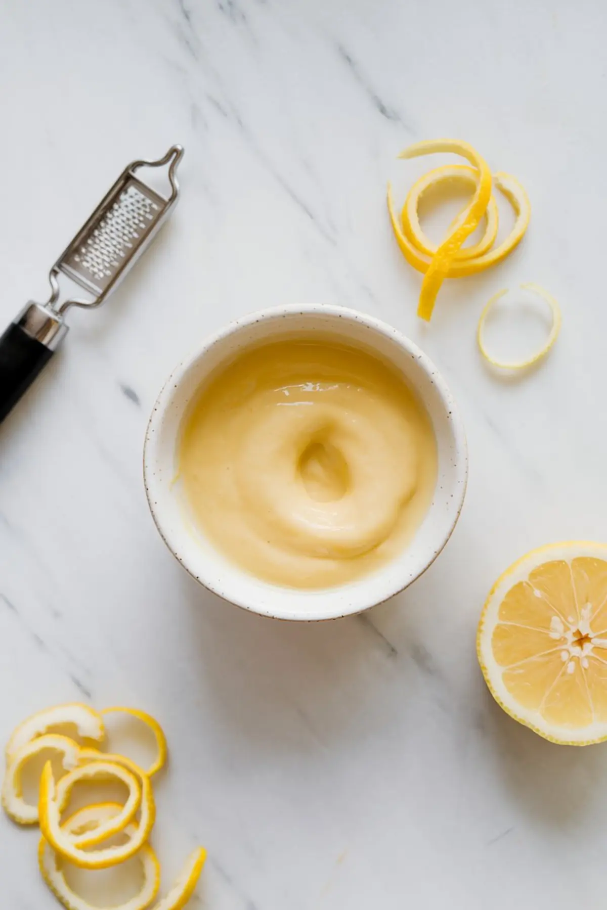 Small bowl of smooth lemon curd surrounded by fresh lemon peel spirals, a grater, and halved lemons, showcasing the tart filling element used in lemon dessert recipes.