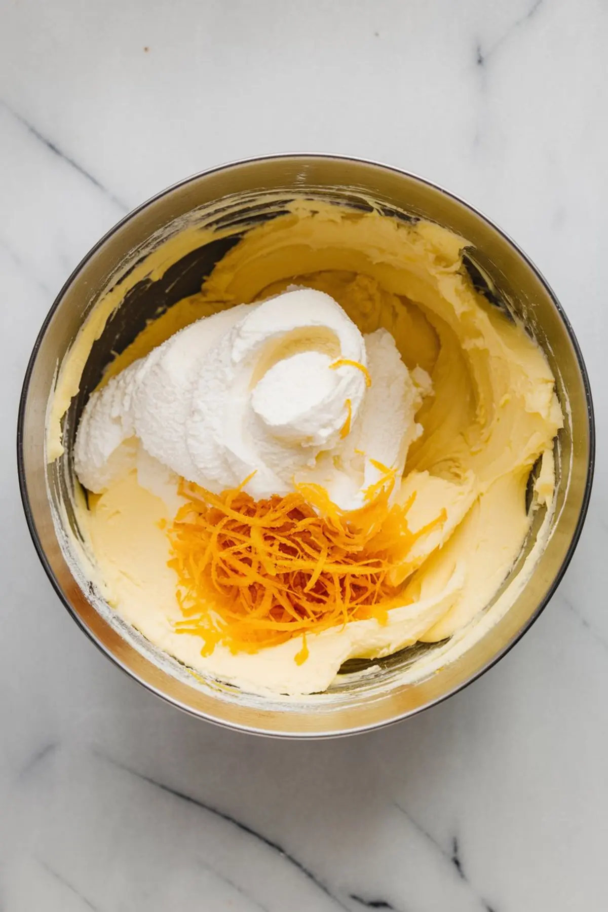 Mixing bowl with whipped butter and powdered sugar topped with vibrant lemon zest, showing a step in preparing citrus cookie dough or frosting base.