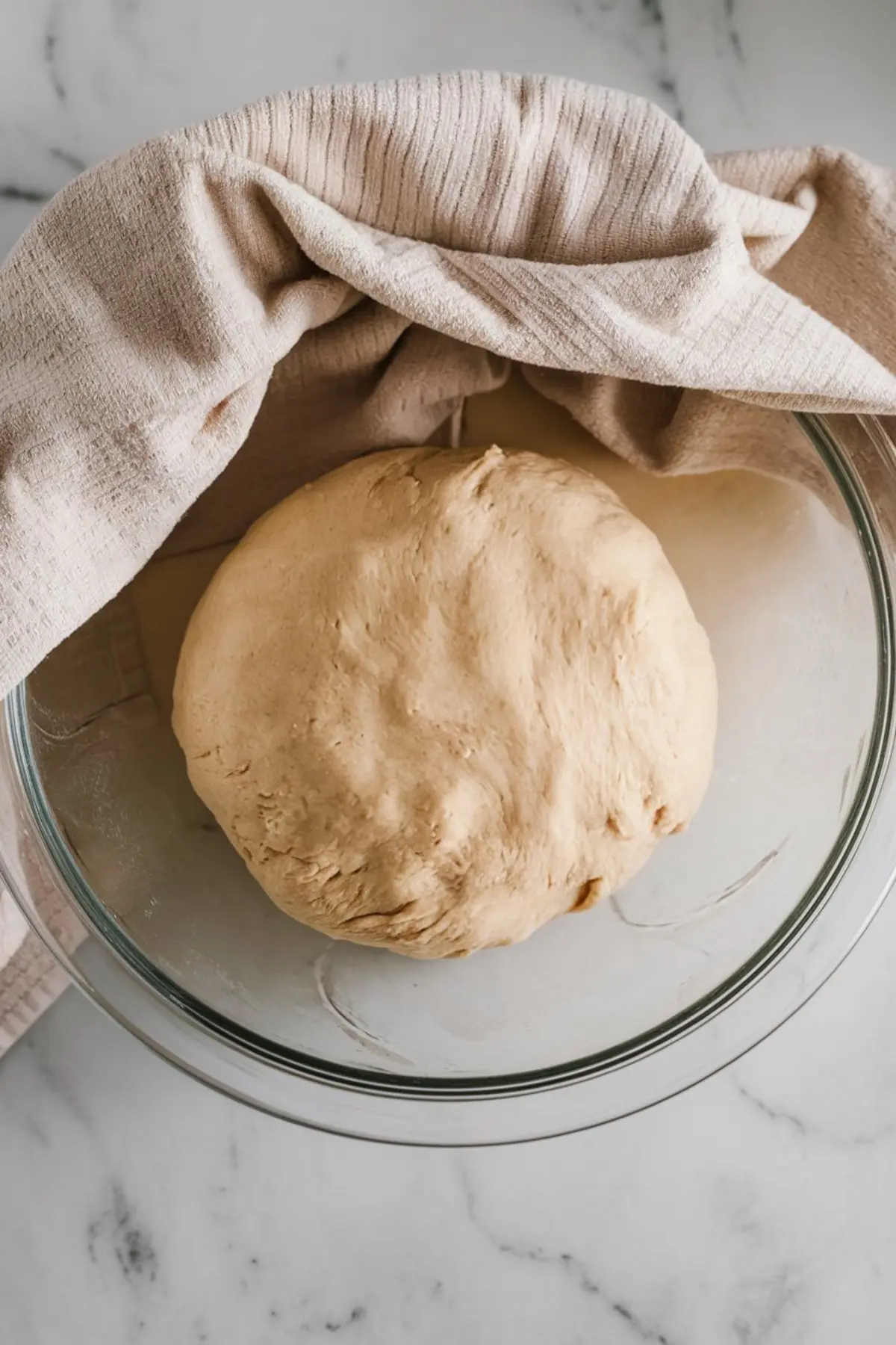 A glass mixing bowl holds a ball of risen dough partially covered with a beige kitchen towel, set on a marble surface, illustrating a proofing stage in homemade bread preparation.