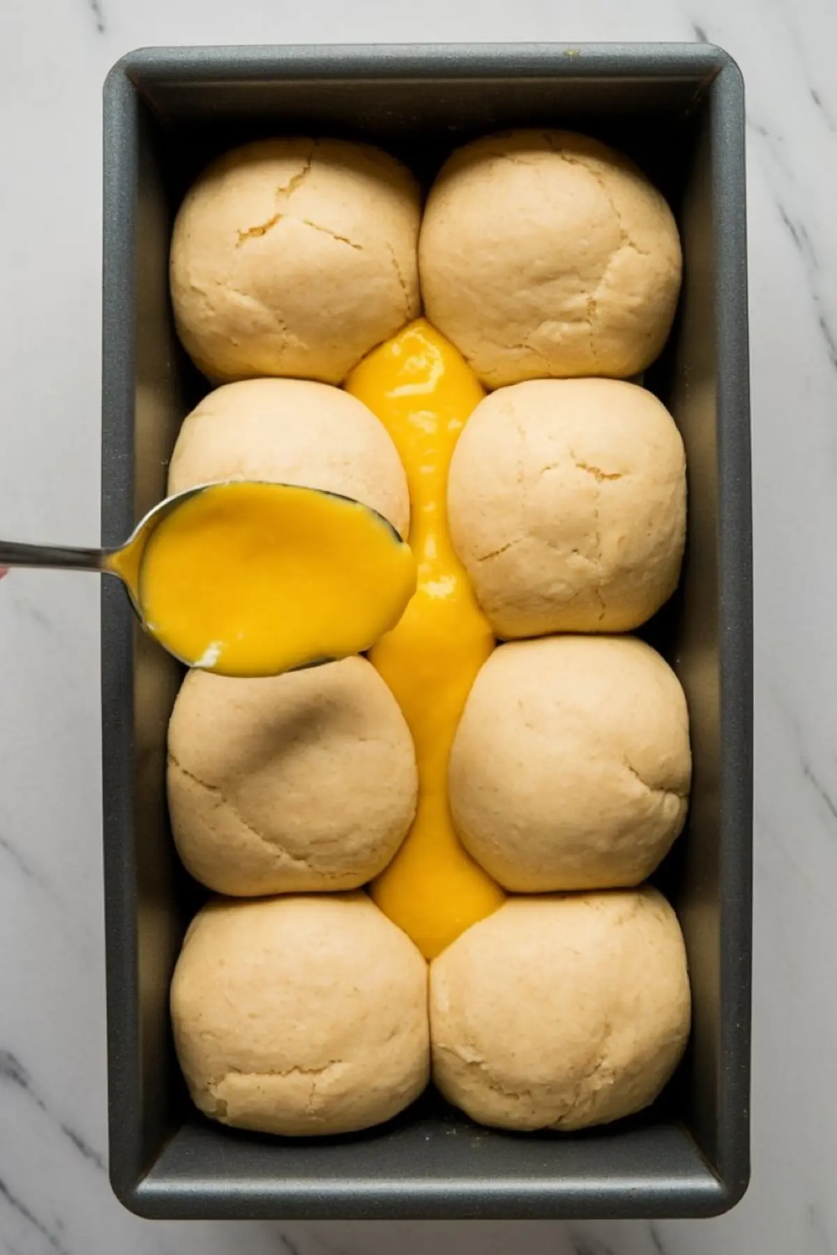 A spoon drizzles vibrant yellow lemon curd into the center of eight dough balls arranged in a loaf pan on a marble surface, capturing a baking preparation step for lemon milk bread.