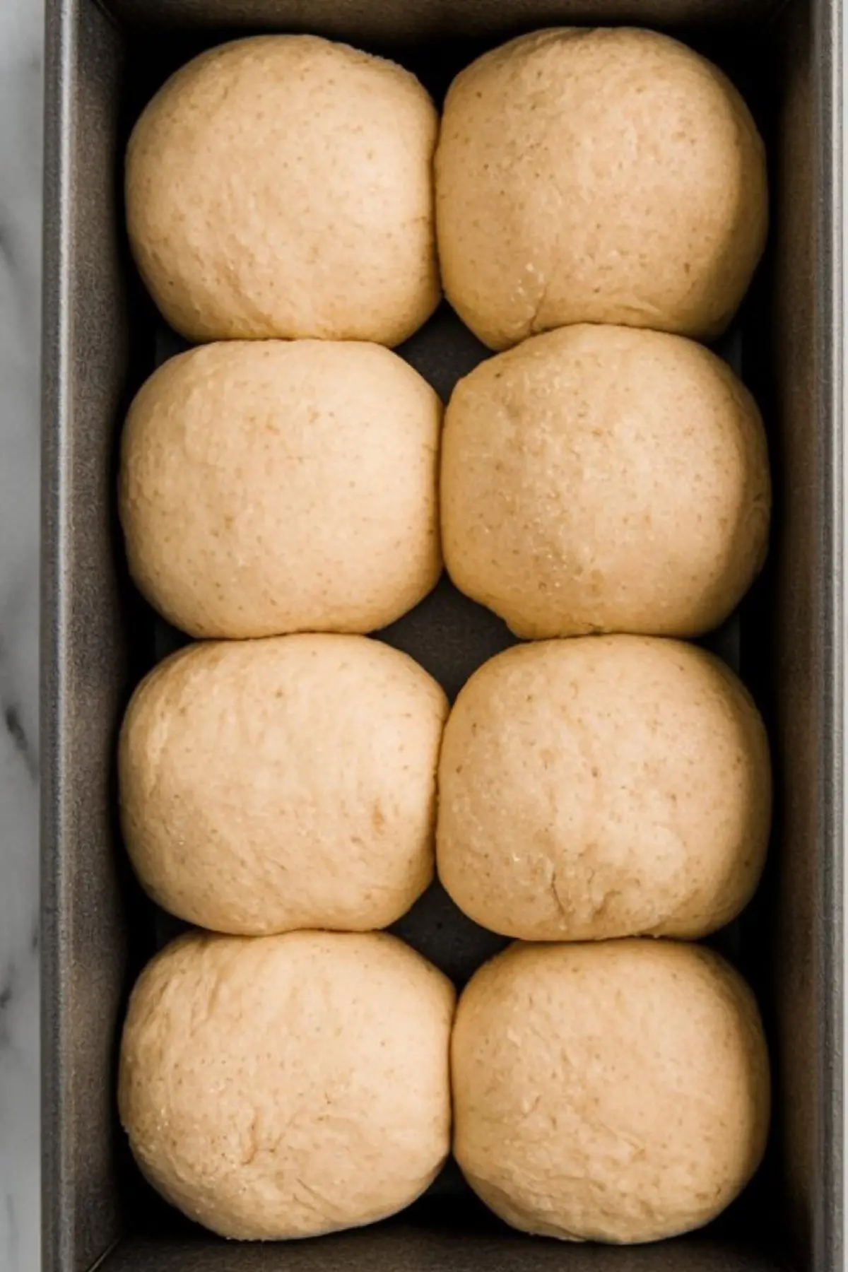 Eight evenly spaced dough balls sit in a loaf pan before baking, set against a white marble background, showcasing an early step in making homemade milk bread.