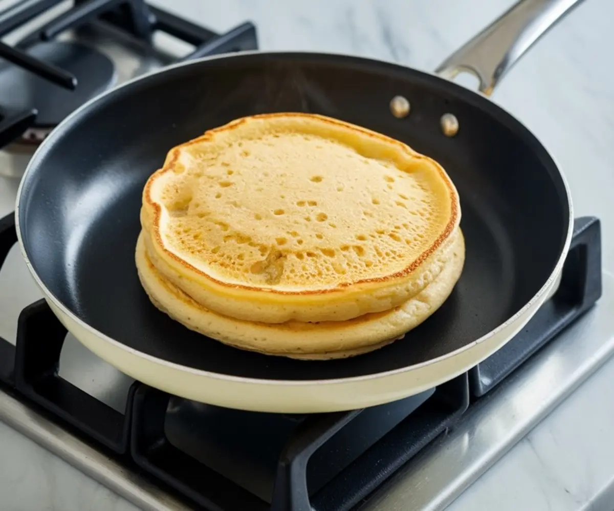 Lemon soufflé pancake cooking in a nonstick skillet on a stovetop, showing golden-brown edges and light, airy texture in the center.