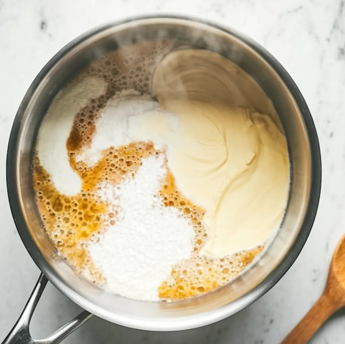 Cream mixture with sugar, cream cheese, and vanilla in a saucepan over heat, showing the cooking stage of mango panna cotta preparation.