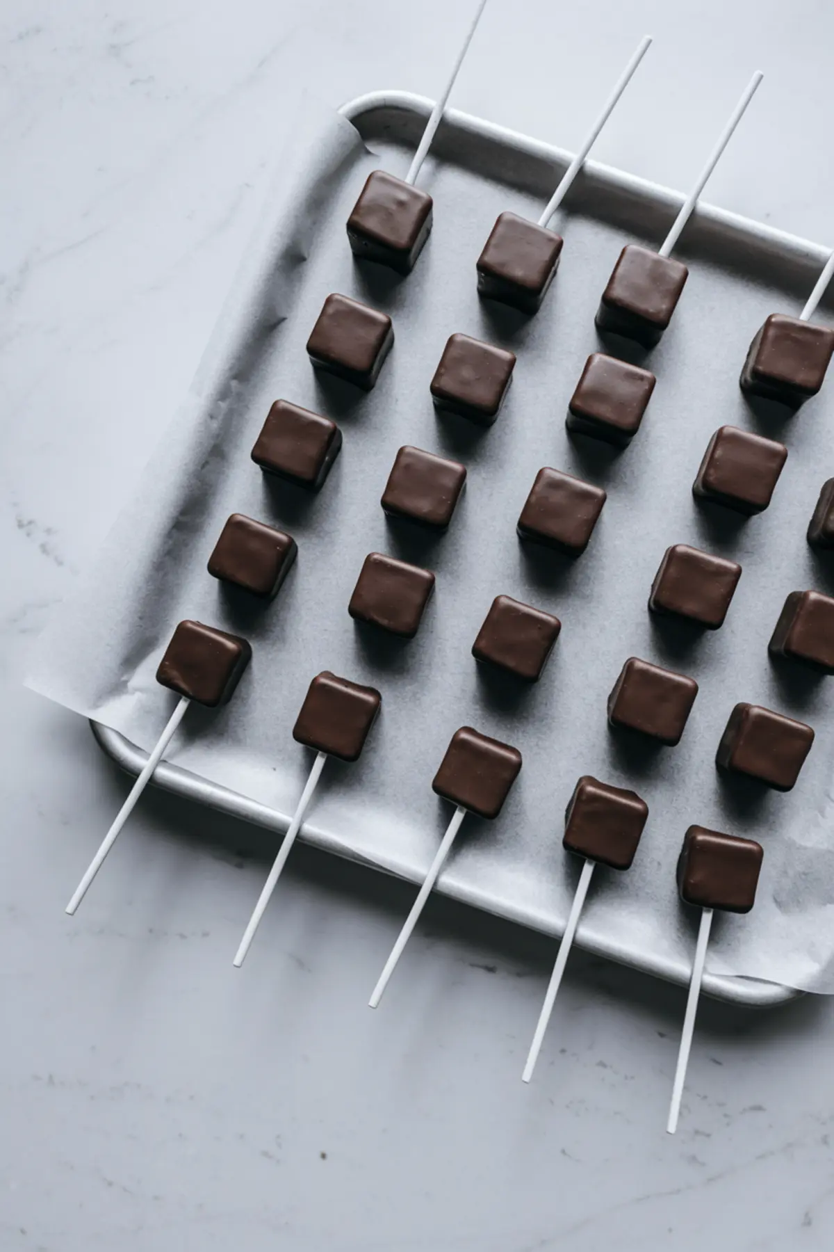 Rows of plain chocolate-coated square cake pops set on white sticks, lined neatly on a parchment-covered tray, ready for decorating.
