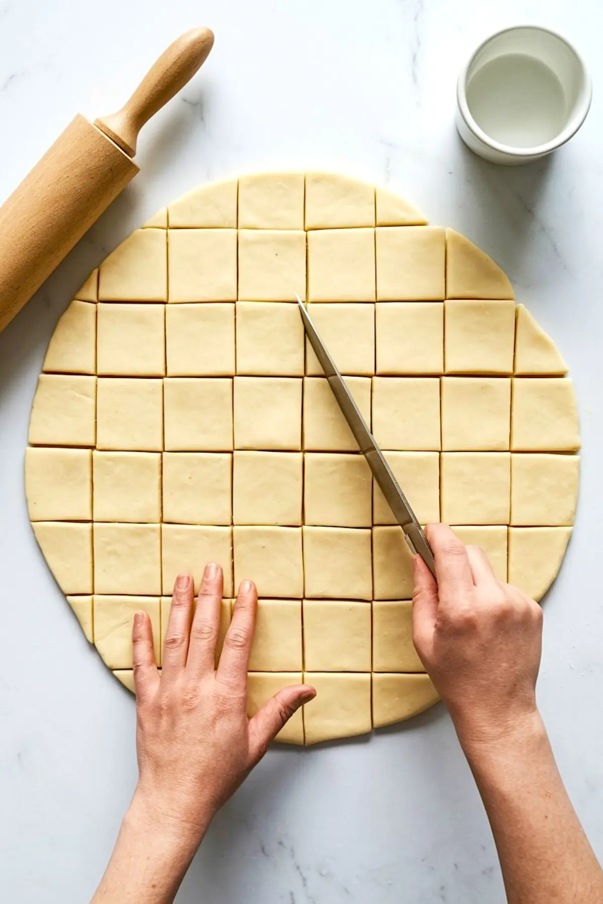 Person slicing rolled-out beignet dough into perfect square pieces with a knife, alongside a wooden rolling pin and small bowl on a marble countertop.