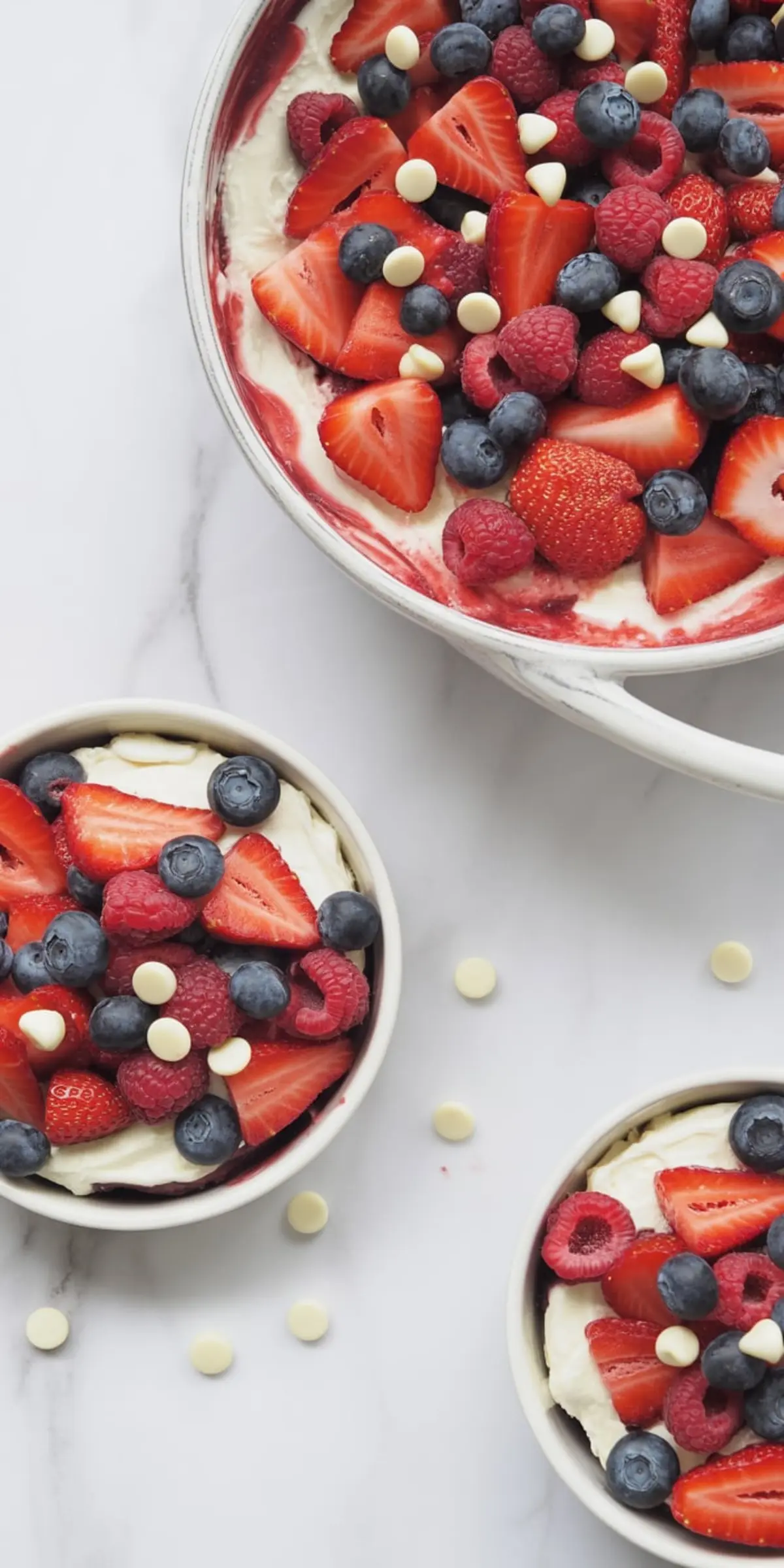 Overhead shot of a large dish and three small bowls filled with berry cheesecake salad, each topped with fresh strawberries, raspberries, blueberries, and white chocolate chips, arranged on a white marble surface.