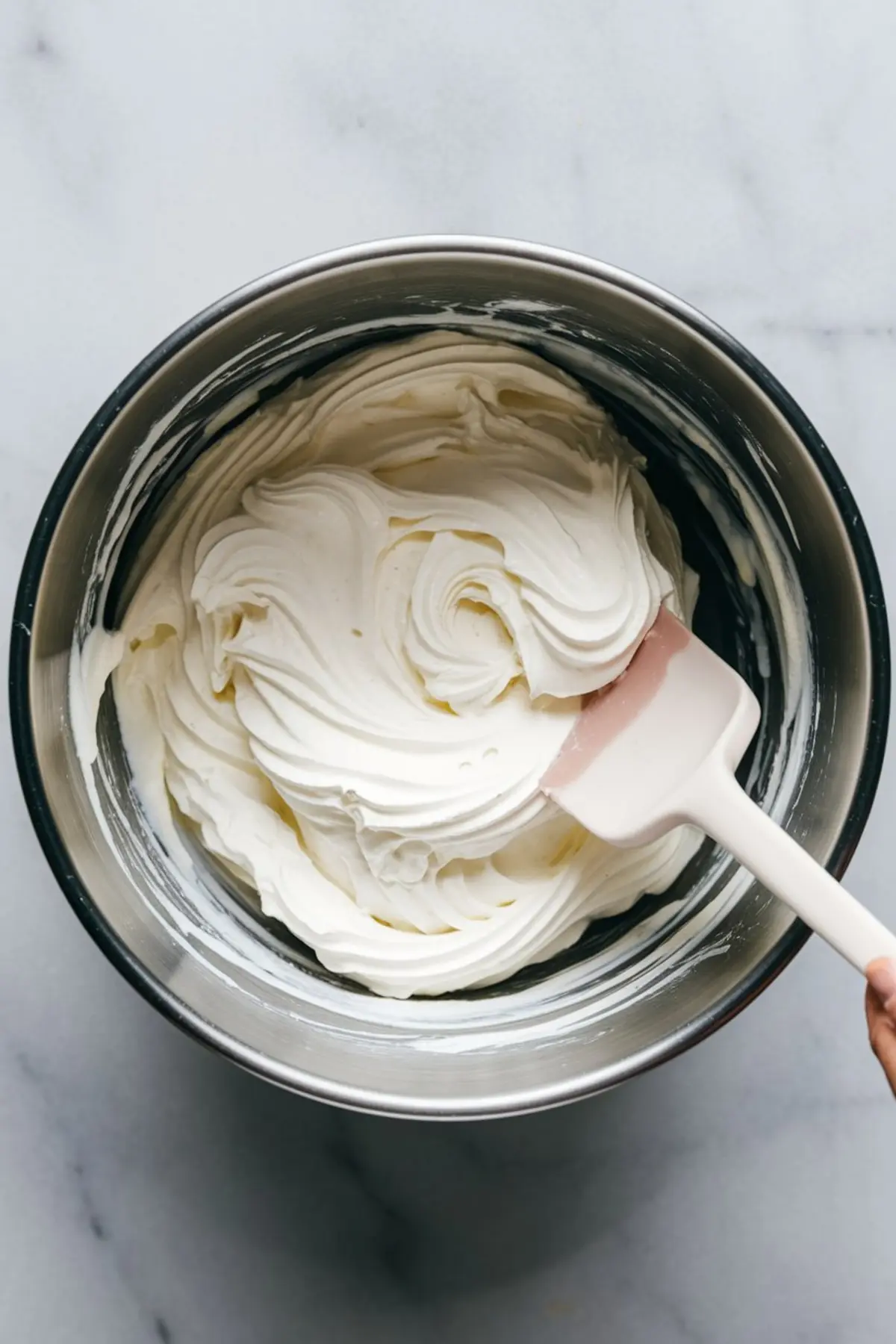 Metal mixing bowl filled with freshly whipped cream cheese mixture, stirred with a white and pink spatula, placed on a white marble countertop.