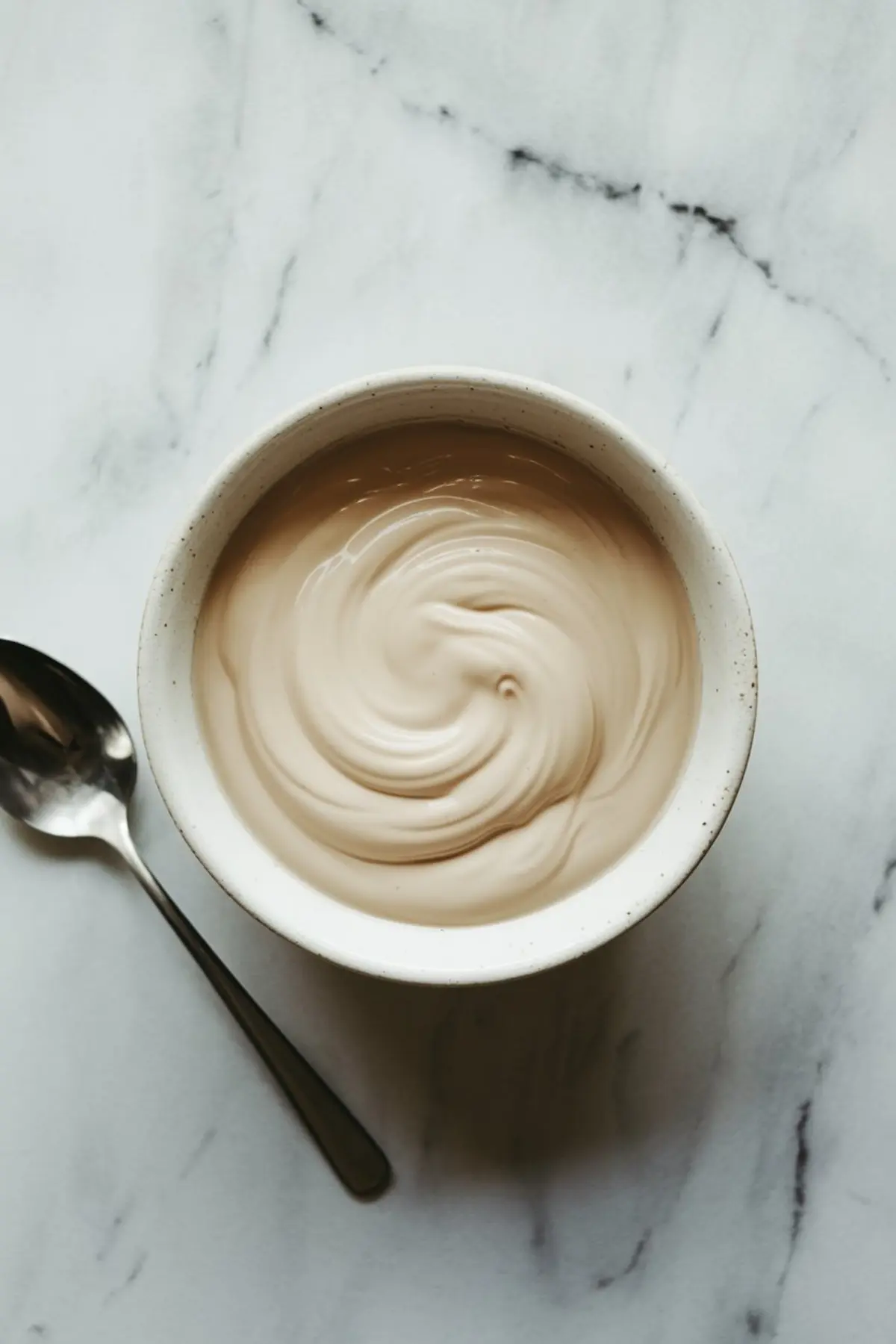 Creamy beige cake batter or frosting swirled in a white ceramic bowl with a metal spoon on a light marble countertop.