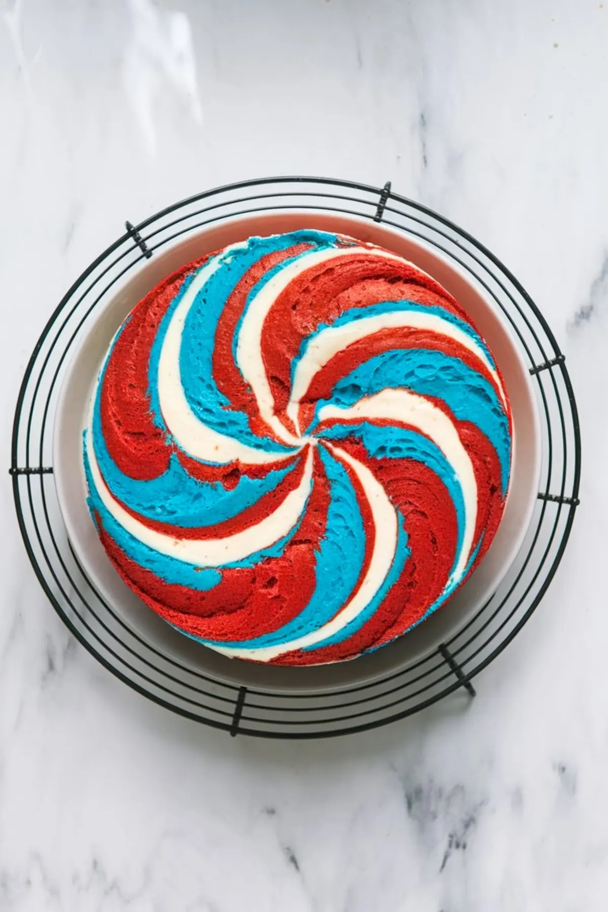 Round baked cake with red, white, and blue swirled layers forming a spiral pattern, cooling on a wire rack over a white marble background.