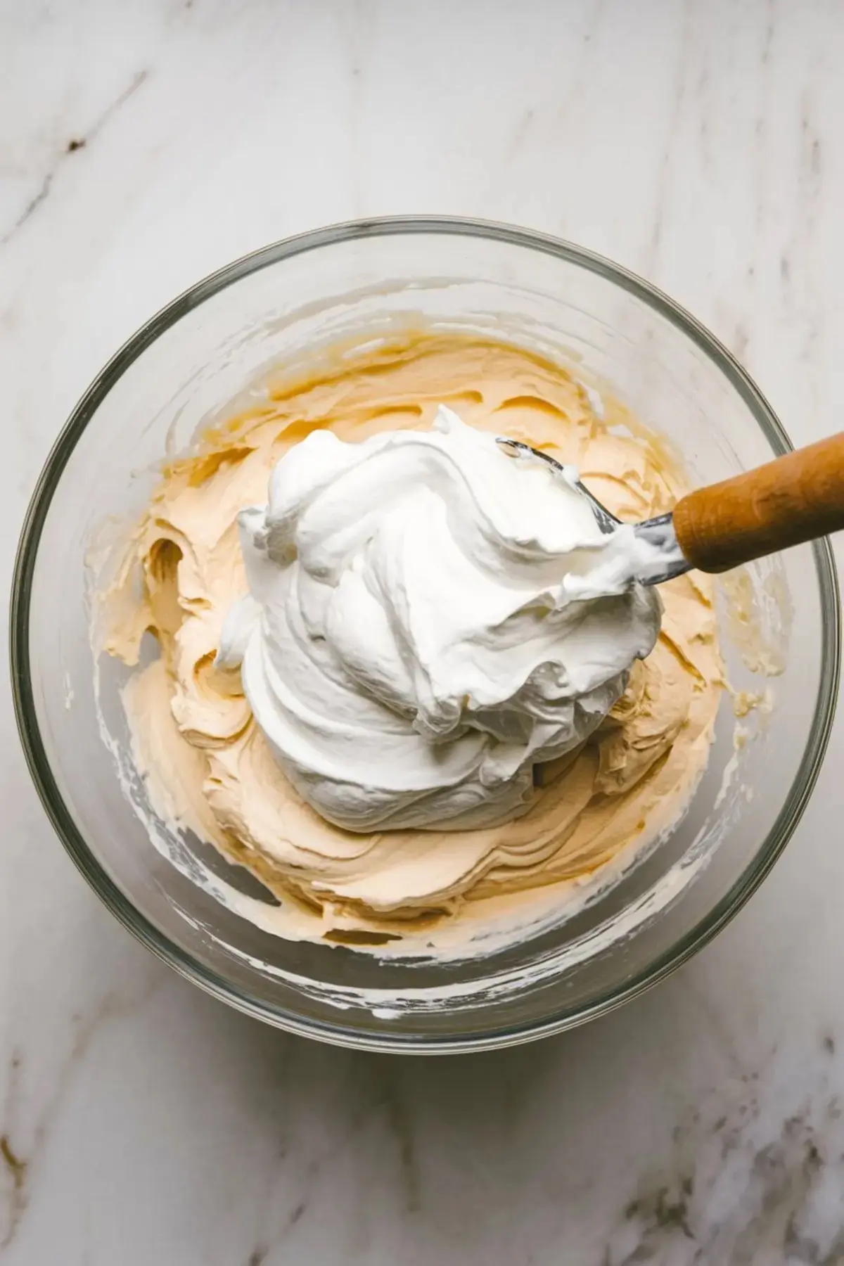 Cheesecake batter with whipped topping folded in for a smooth, airy texture, shown in a clear mixing bowl on marble.