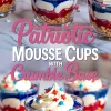 Red, white, and blue mousse cups with crunchy crumble base, each topped with whipped cream and star sprinkles, arranged on a marble surface with scattered decorative sprinkles.