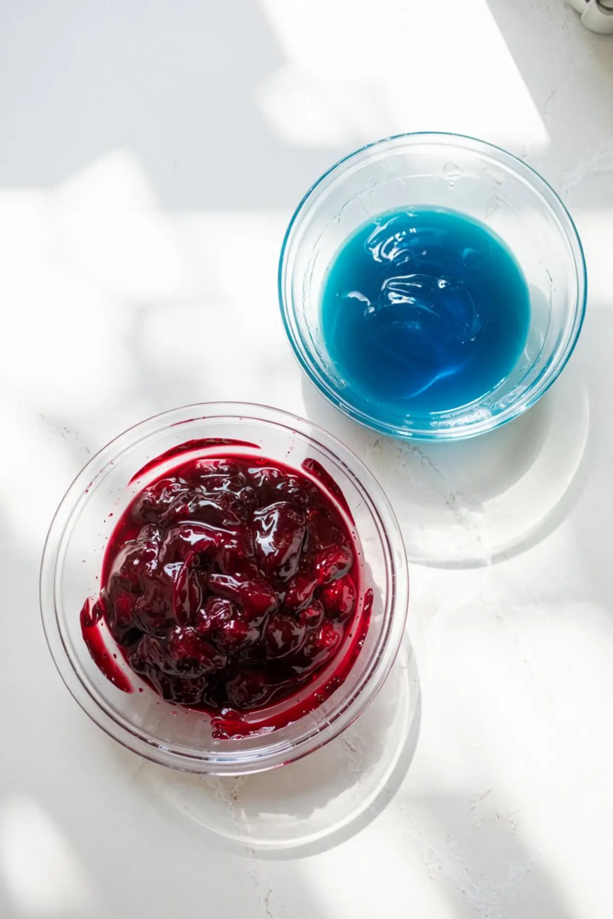 Glass dessert bowls filled with deep red cherry pie filling and vibrant blue gelatin, placed side by side on a sunlit white counter as mousse ingredients.