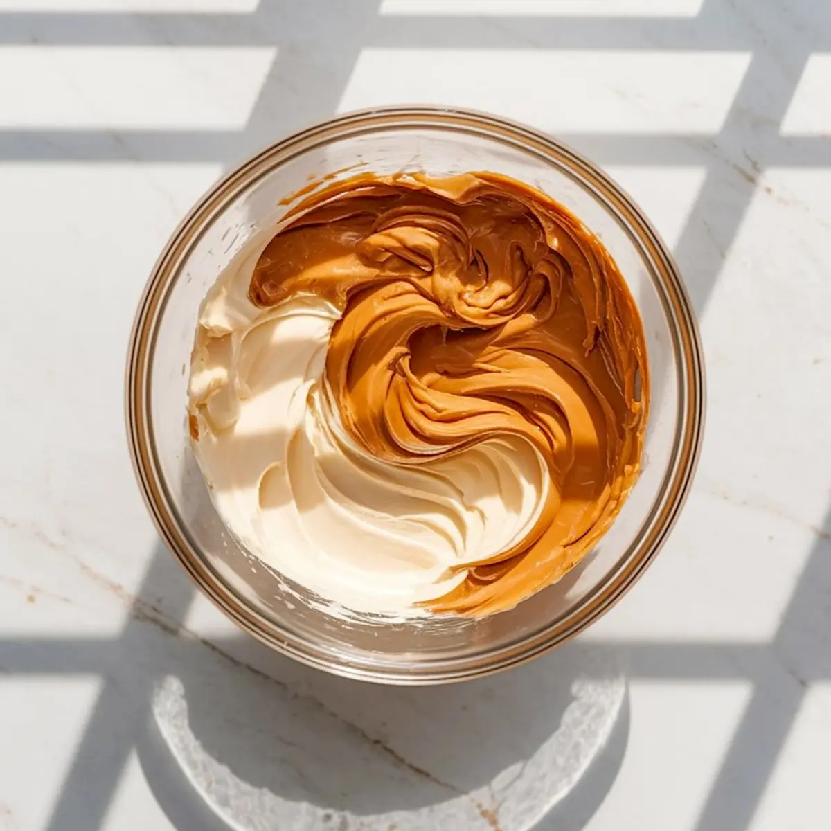 Swirled mixture of peanut butter and cream cheese in a glass bowl, ready to be blended for cheesecake batter preparation.