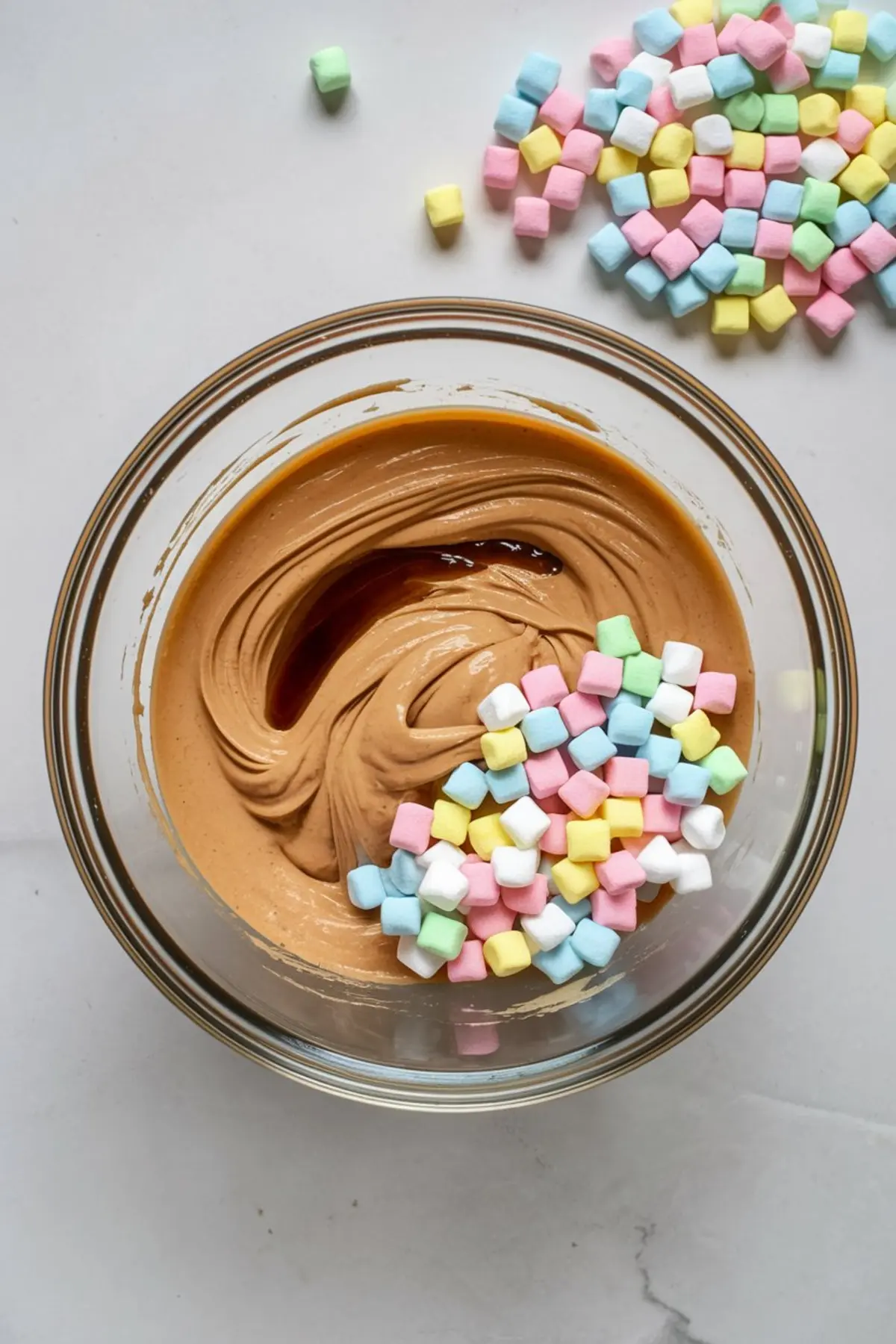 Glass mixing bowl with swirled peanut butter, vanilla extract, and pastel mini marshmallows, with extra marshmallows scattered on a white surface nearby.