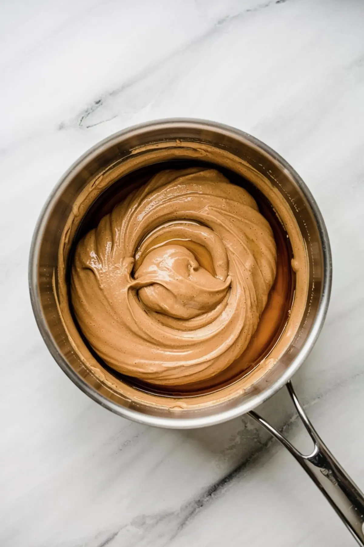 Creamy peanut butter mixture swirled smoothly inside a stainless steel saucepan, prepared on a white marble countertop for no-bake cookie filling or dessert base.