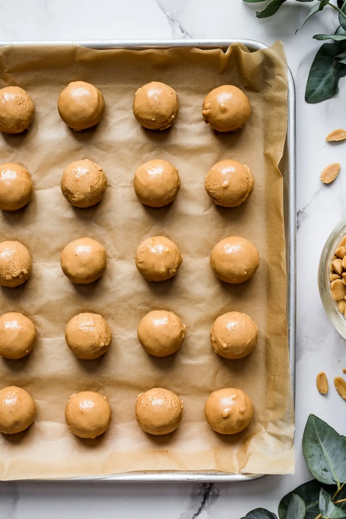 Round peanut butter cookie dough balls lined on a baking sheet with parchment paper, ready for chilling or coating, viewed from above on a marble surface with scattered peanuts and green leaves.