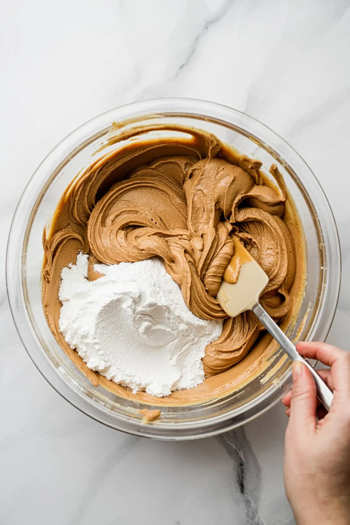 Thick peanut butter cookie dough mixture being folded with powdered sugar in a large glass bowl using a silicone spatula, seen from above on a white marble background.