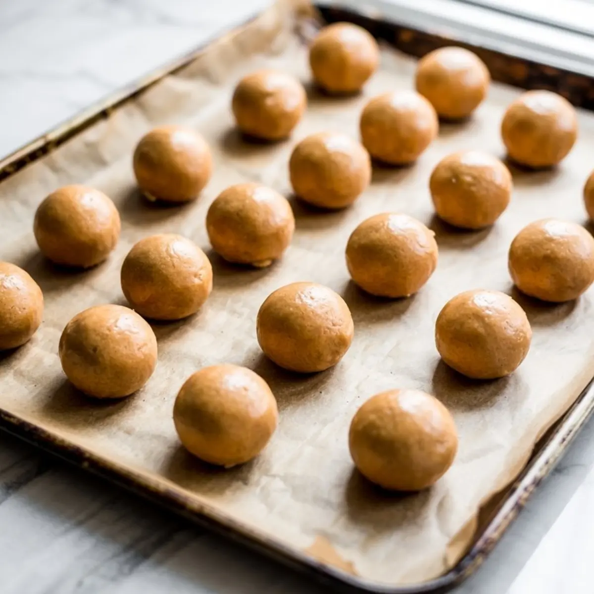 Rows of evenly shaped peanut butter cookie dough balls arranged on parchment-lined baking tray, prepared for a no-bake dessert recipe.