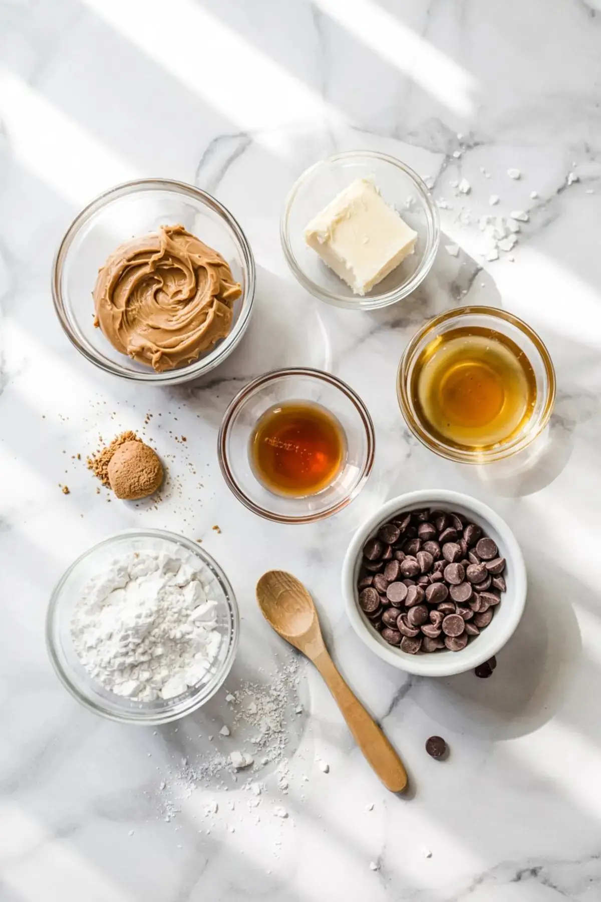 Overhead view of no-bake peanut butter cookie ingredients in glass bowls, including creamy peanut butter, powdered sugar, chocolate chips, maple syrup, butter, and vanilla extract on a white marble counter.