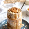 Mason jar filled with layered peanut butter overnight oats, oats on top with a swirl of peanut butter, and text overlay reading “peanut butter overnight oats.” Placed on a blue plate with oats scattered. Great for over night oats in a jar and protein overnight oats healthy content.