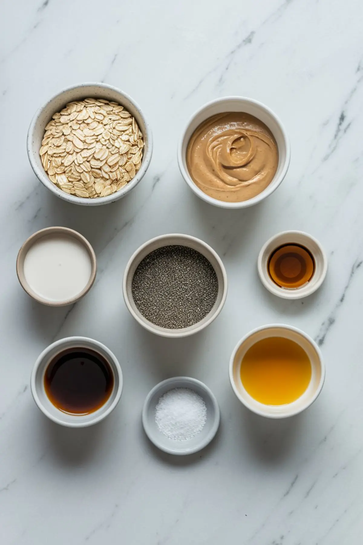 Flat lay of overnight oats ingredients on a marble surface, including rolled oats, peanut butter, chia seeds, plant-based milk, vanilla extract, maple syrup, sea salt, and melted coconut oil. Ideal for prepping over night oats in a jar or blended overnight oats.