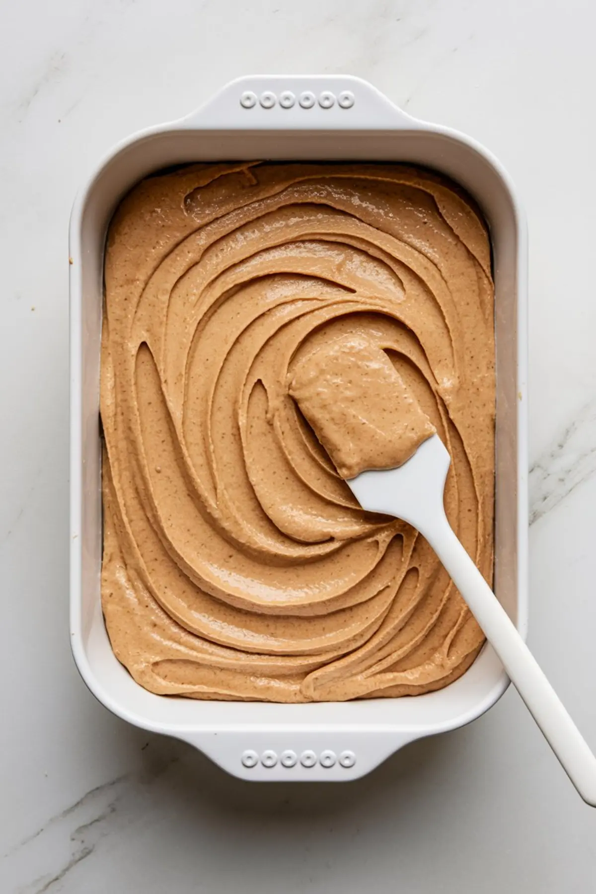 Top view of peanut butter cake batter evenly spread in a white baking dish with a spatula, ready for baking, showcasing smooth, swirled surface texture.
