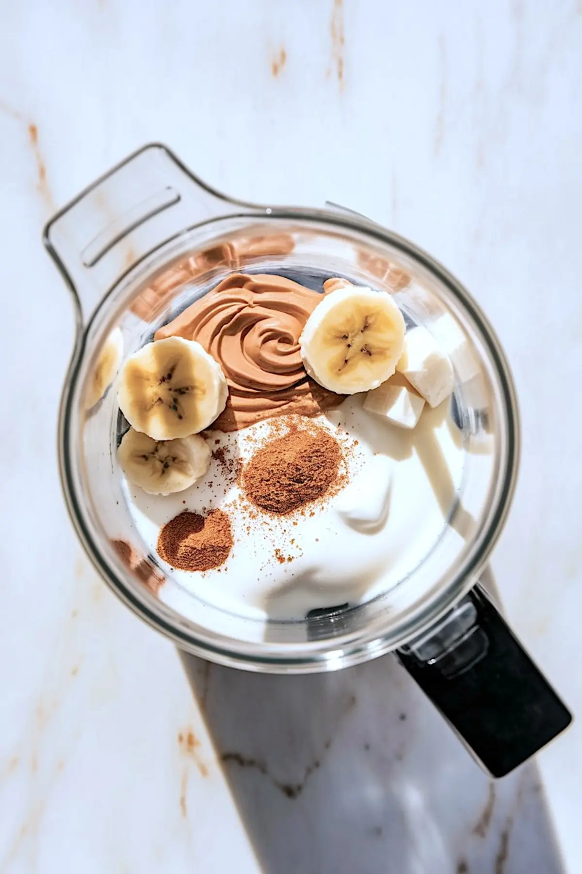 Top view of a blender filled with sliced bananas, creamy peanut butter, Greek yogurt, cubes of ice, and a sprinkle of cinnamon, ready to blend into a protein-packed peanut butter smoothie.