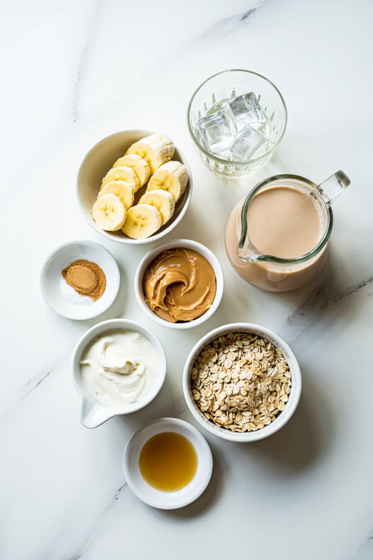 Flat lay of peanut butter smoothie ingredients including sliced bananas, rolled oats, yogurt, peanut butter, ice cubes, cinnamon, honey, and a glass of oat milk, arranged on a white marble surface.