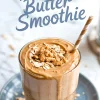 Thick peanut butter smoothie in a clear glass topped with granola and oats, set on a gray plate with a striped straw, captioned “Peanut Butter Smoothie” for smoothie aesthetic inspiration.