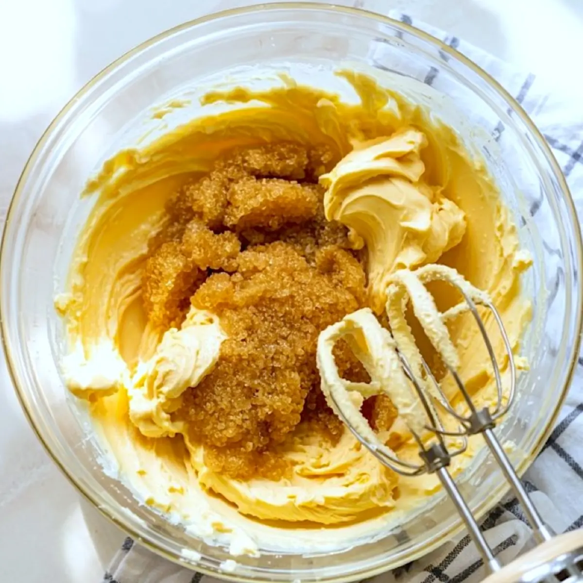 Mixing bowl filled with creamy butter and brown sugar being blended with an electric mixer, showing the early stage of cookie dough preparation on a white checkered towel.
