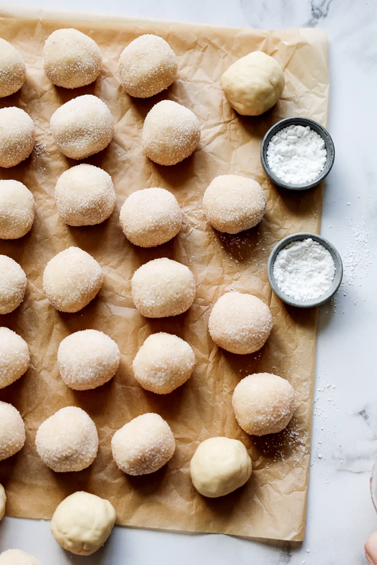 Rows of unbaked cookie dough balls coated in sugar arranged on a parchment-lined surface, with small bowls of powdered sugar and plain dough in the corner, set against a marble background.