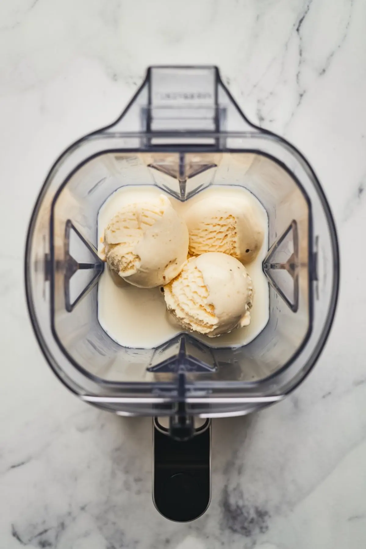 Top-down view of a blender containing three scoops of vanilla ice cream and milk, ready to be blended for a creamy dessert shake.